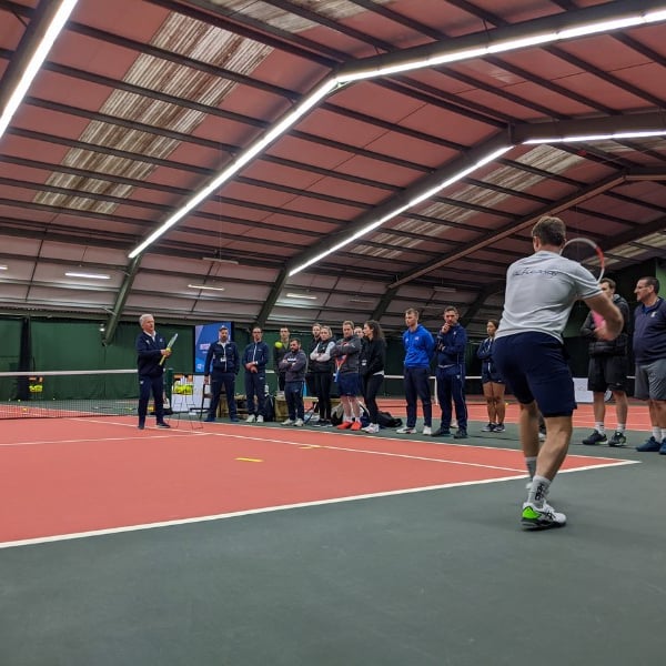 Indoor tennis training at David Lloyd West Bridgford with players watching a coach demonstrate a drill.