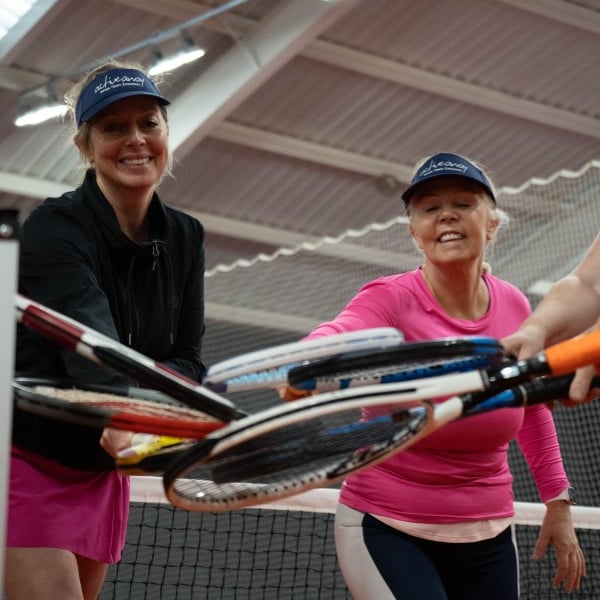 Three women in tennis outfits with visors, joining their racquets together inside an indoor tennis court, expressing teamwork and camaraderie.