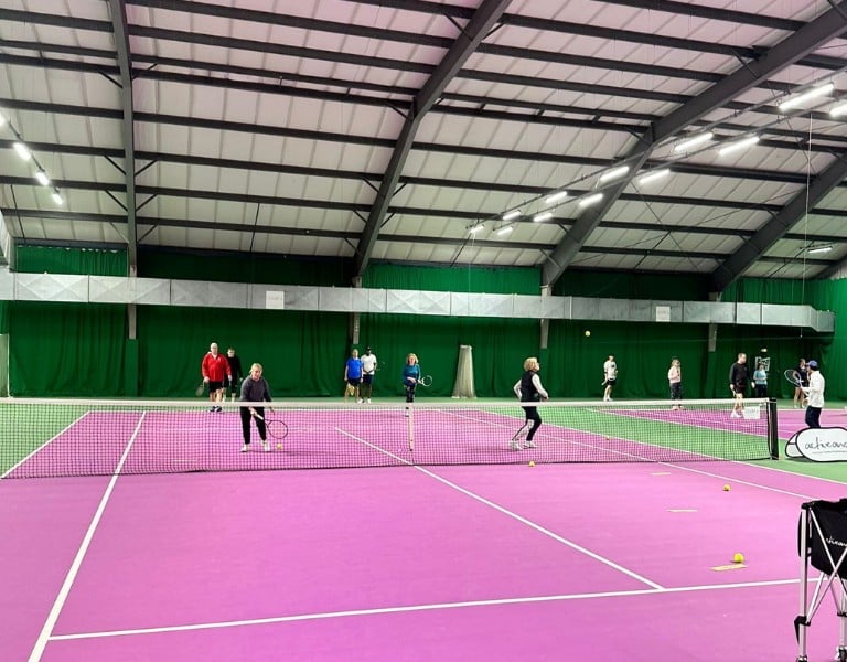 Indoor tennis court in Woking with players engaged in a doubles match on a pink surface and green walls.