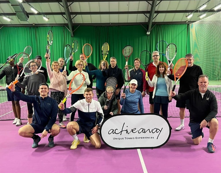 Group of tennis players at DL Woking holding rackets and a banner for Active Away Unique Tennis Experiences on an indoor court.