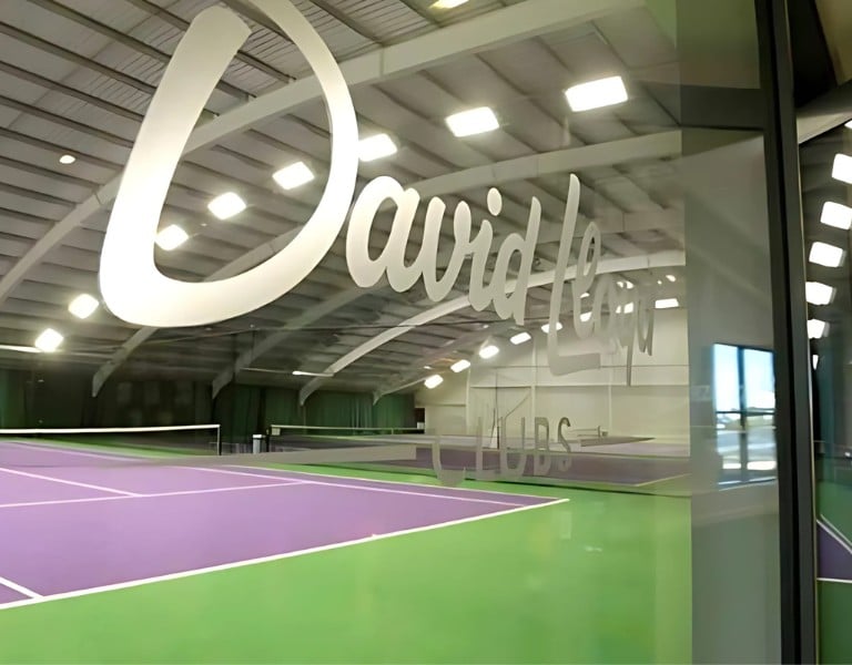 Indoor tennis court at David Lloyd Clubs, Woking, with purple and green floor and David Lloyd Clubs logo on glass window.