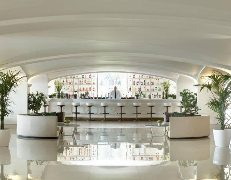 Daphnila Bay Bar with a modern white design, curved ceilings, well-stocked shelves, bar stools, and potted plants.