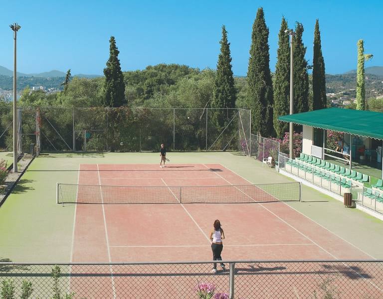 Tennis court at Daphnila Bay with two players, surrounded by greenery and spectator seating, under a clear blue sky.