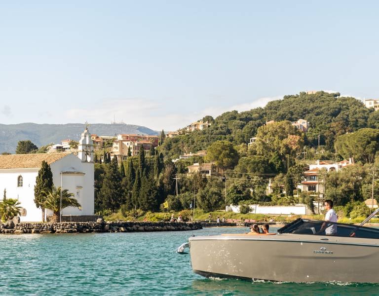View of a cruise in Daphnila Bay with a backdrop of green hills, buildings, and a white chapel by the water.