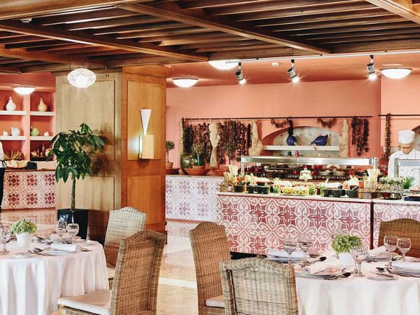 Indoor view of Daphnila Bay restaurant, with round tables, wicker chairs, and a buffet station in a warmly decorated setting.