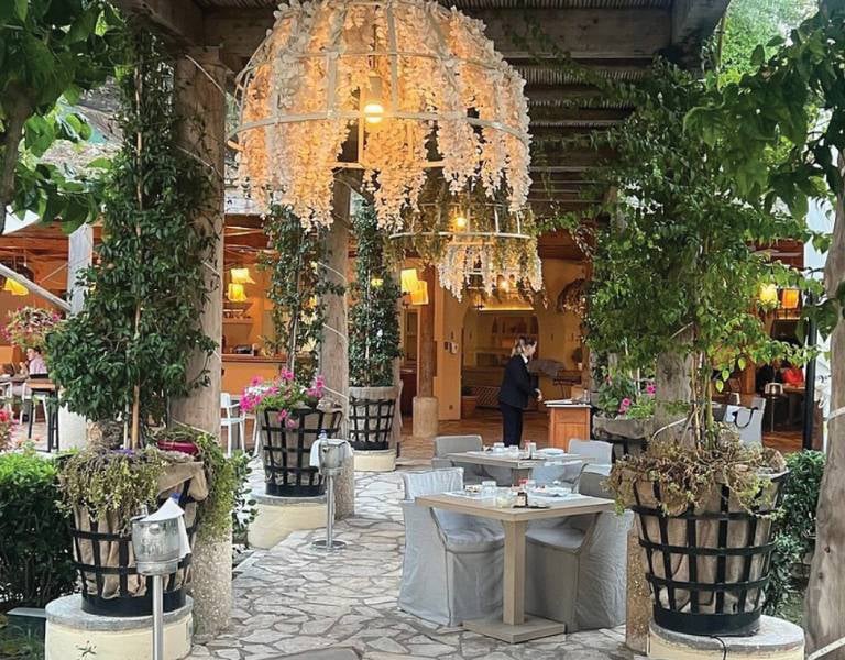 Outdoor restaurant at Daphnila Bay with rustic wood beams, hanging white flower chandeliers, lush greenery, and stone pathway.