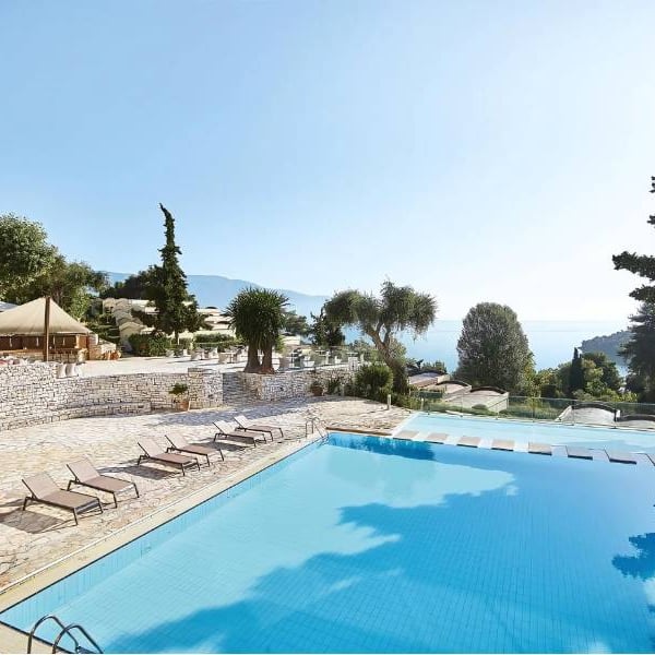 Swimming pool at Daphnila Bay surrounded by lounge chairs and greenery, overlooking the sea and a clear sky.