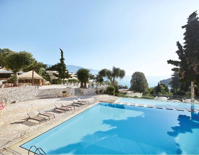 Swimming pool at Daphnila Bay surrounded by lounge chairs and greenery, overlooking the sea and a clear sky.
