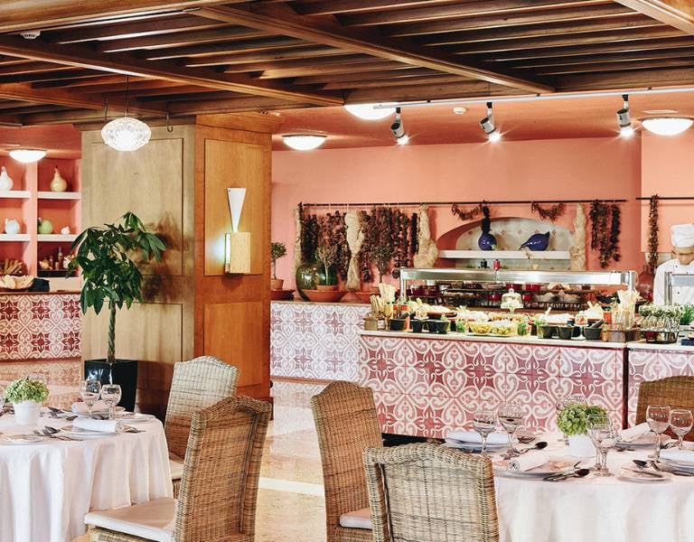 Restaurant interior at Daphnila Bay with set tables, wicker chairs, and a buffet with decorative tiles and food options.