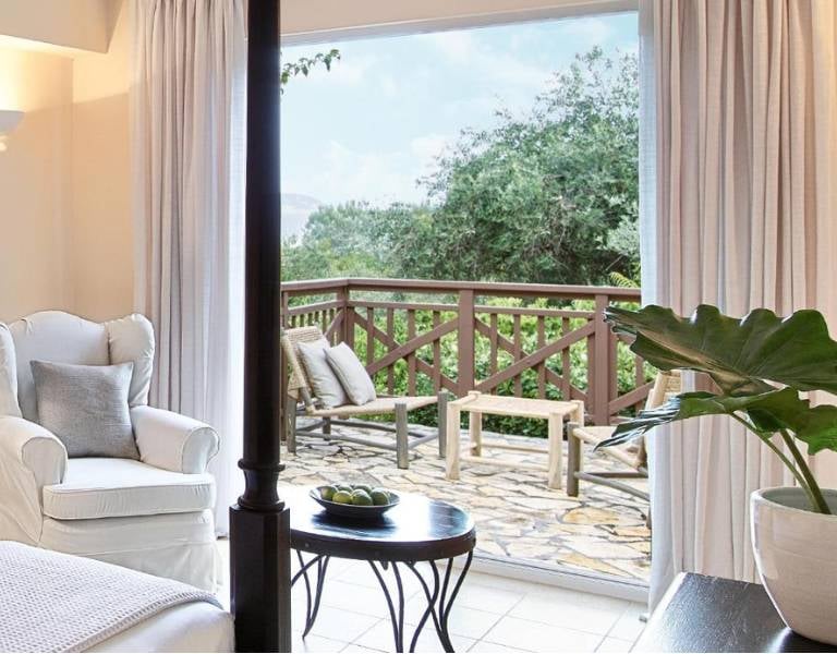 Balcony view from a room at Daphnila Bay, with a white armchair, round table, and lush greenery outside.