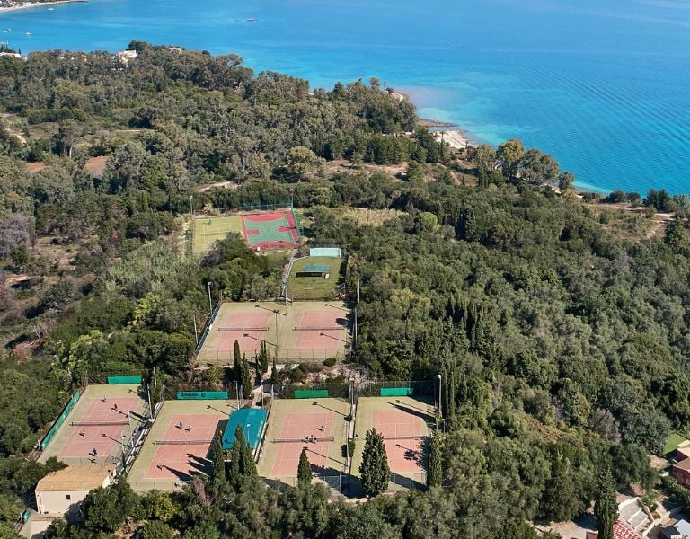 Aerial view of tennis courts at Daphnila Bay, surrounded by greenery and overlooking blue waters in Corfu, Greece.