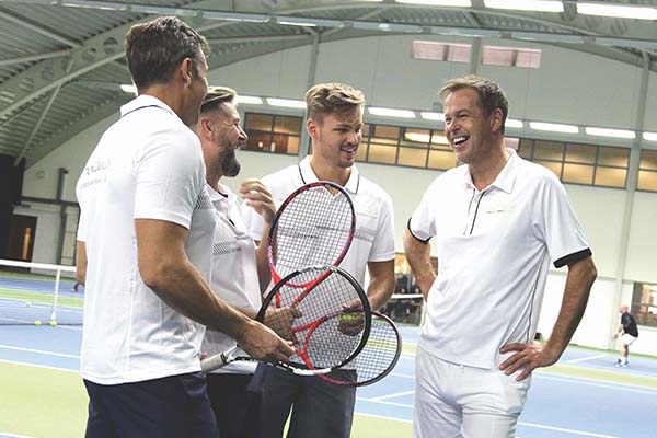 Josh Thompson and three other men with tennis rackets on an indoor tennis court, laughing and chatting in white sportswear.