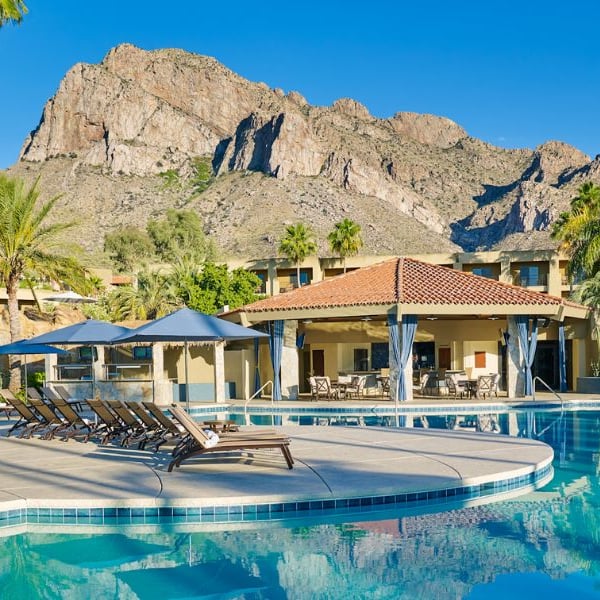 Pool area at El Conquistador Resort with lounge chairs, parasols, and mountainous backdrop under a clear sky.