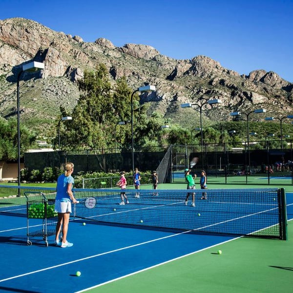 Outdoor tennis match at El Conquistador with players and coach, set against a backdrop of mountains and blue sky.