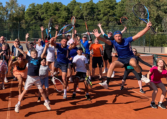 Families and friends celebrating on a red clay tennis court, holding rackets and cheering during a family tennis holiday.