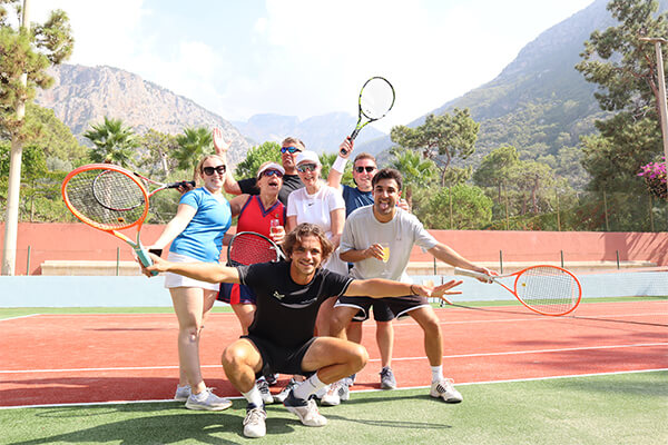 Group of six people smiling and holding tennis rackets on an outdoor court in Turkey with mountains in the background.