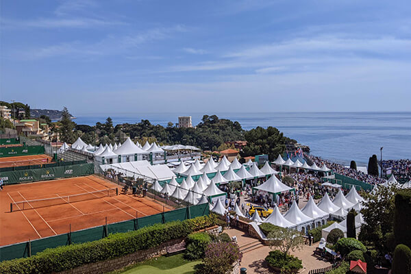 Outdoor grand slam tennis event with clay courts and white hospitality tents by the seaside.