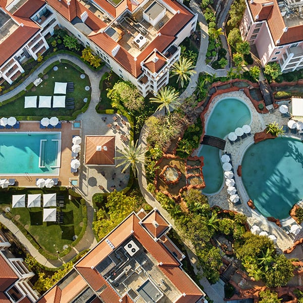 Aerial view of Hilton Vilamoura with two swimming pools and surrounding gardens, featuring sun loungers and red-tiled buildings.