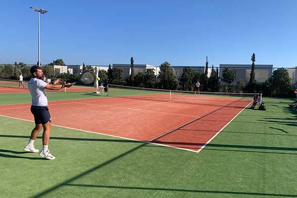 Tennis player in white shirt and navy shorts hitting a forehand on a red clay court, with spectators watching and trees in the background.
