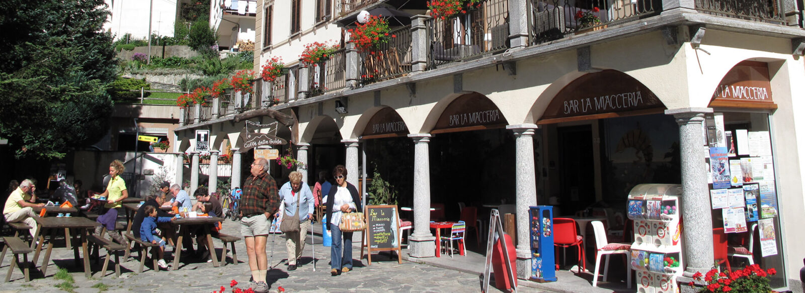 Hotel Monterosa Bar exterior with patrons dining at outdoor tables, stone arches, and red flower accents.