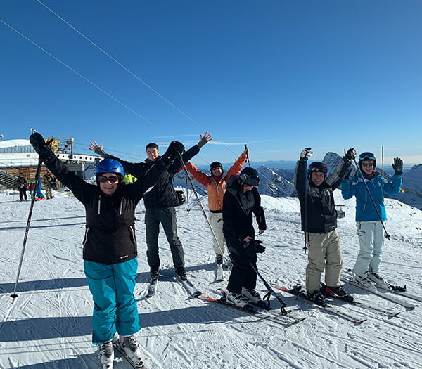 Six skiers on a snowy mountain slope with arms raised, wearing skiing gear against a clear blue sky.
