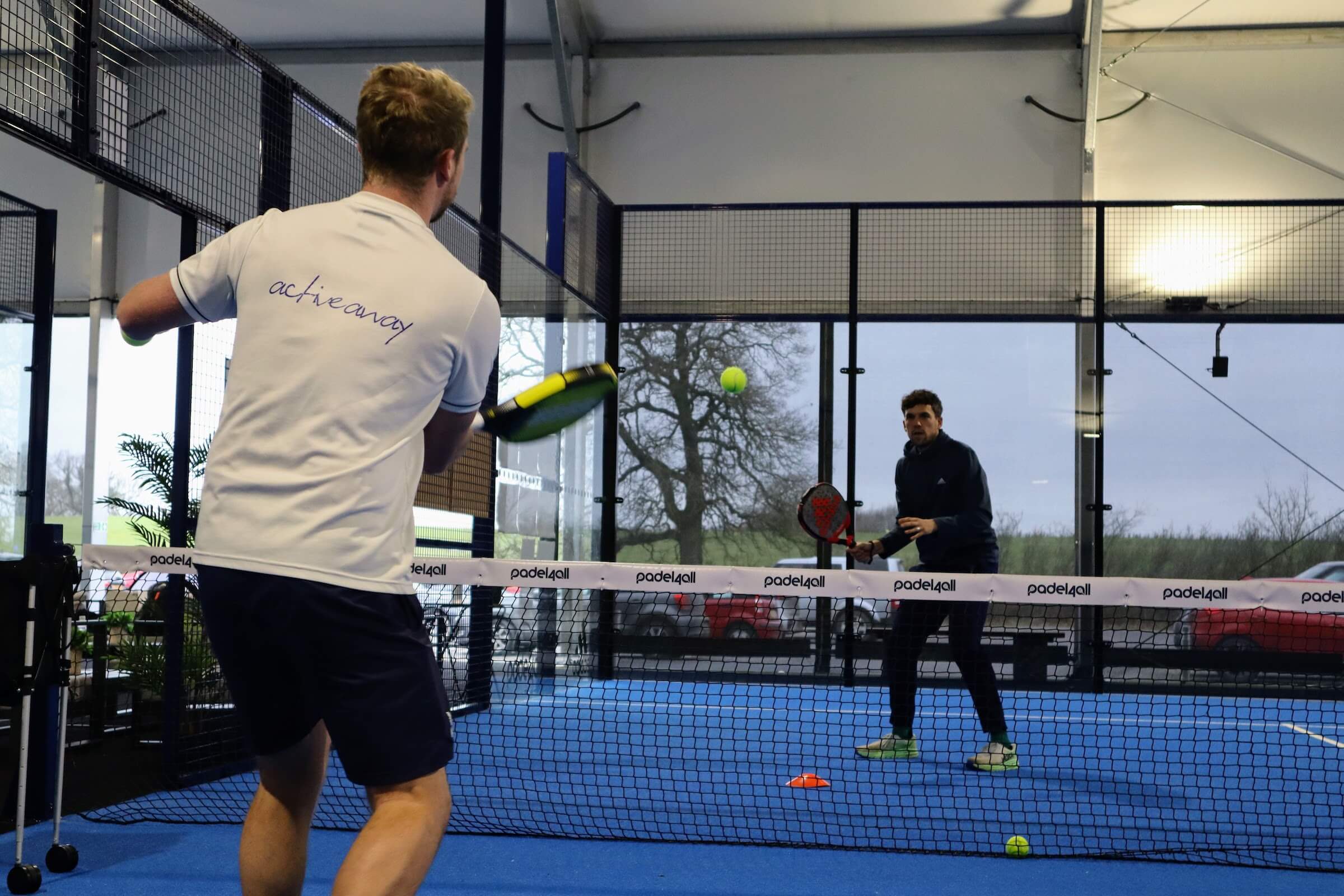 Two men playing padel tennis indoors, with one player winding up for a backhand shot.