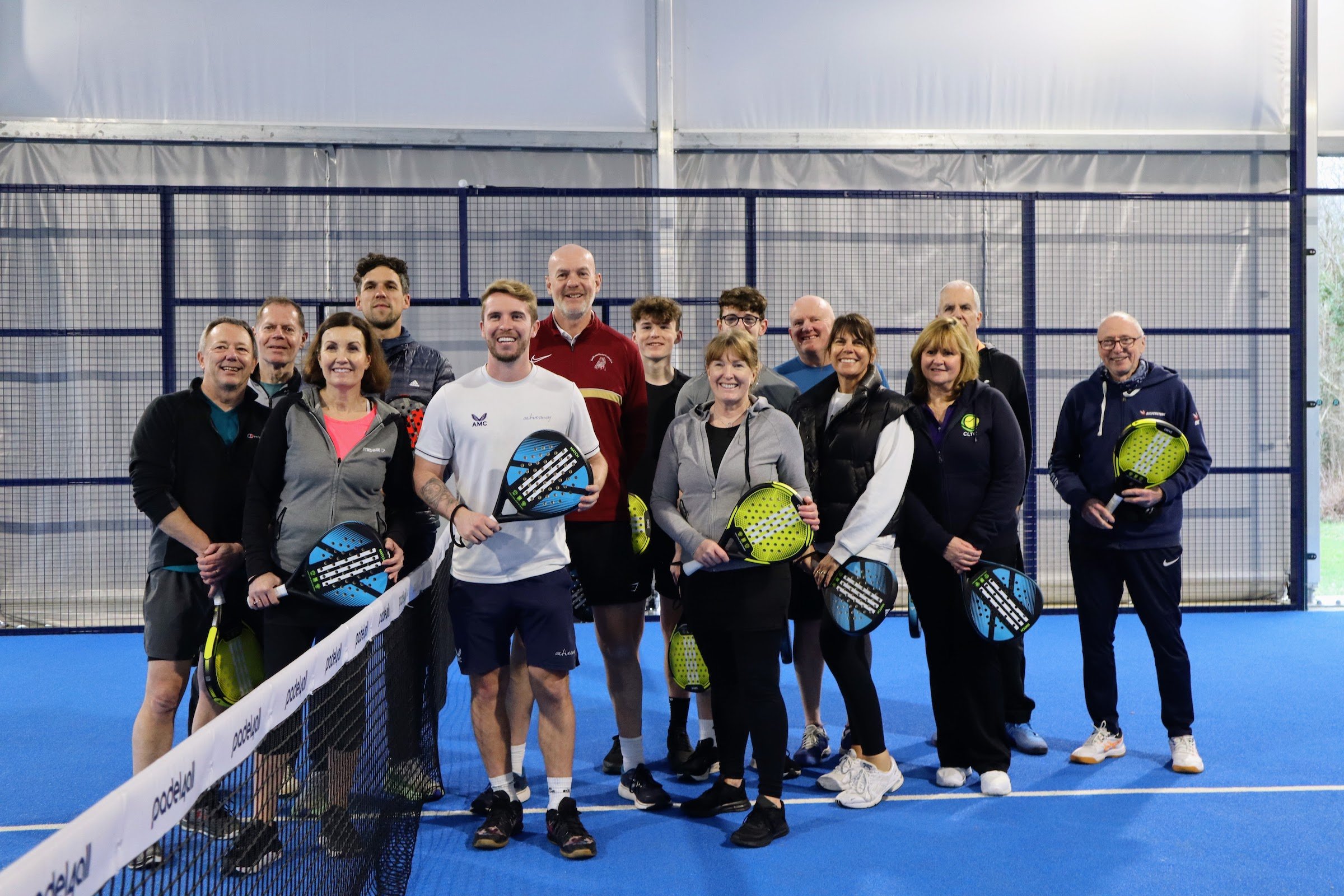 Group of fourteen people holding padel rackets, smiling on a blue court during an indoor padel session.