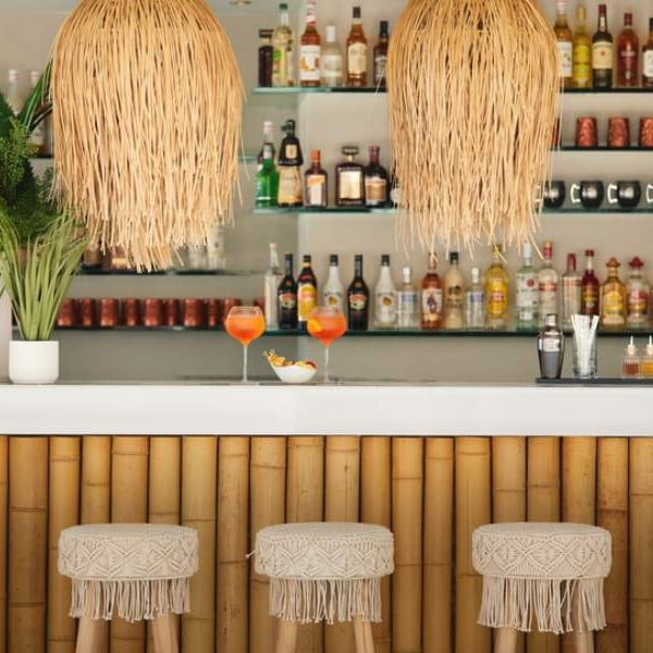 Ikos Aria Gallery bar with bamboo counter, woven stools, rattan pendant lights, and spirits on glass shelves, with two orange cocktails on the counter.
