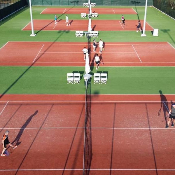 Overhead view of tennis courts at Ikos Aria with players and sunlight casting shadows, surrounded by greenery.
