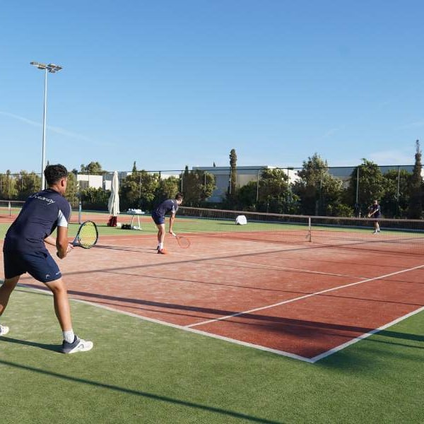 Tennis doubles match in progress on a green and red clay court at Ikos Aria, with players actively engaged under sunny skies.