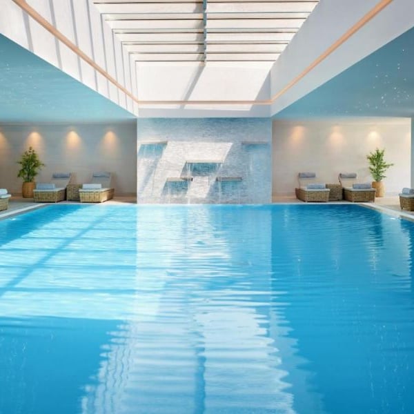Luxurious indoor swimming pool at Ikos Aria with skylight, surrounded by sun loungers and elegant decor.