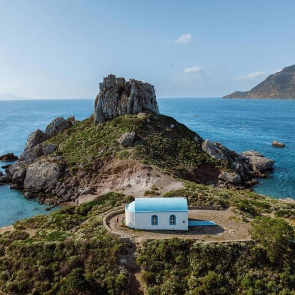 Aerial view of a small white chapel with blue roof on a rocky hill near the Aegean Sea, with cliffs and hills in the background.