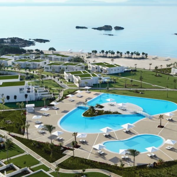 Aerial view of Ikos Aria resort, featuring a large swimming pool, white buildings with green roofs, and proximity to a sandy beach and ocean.