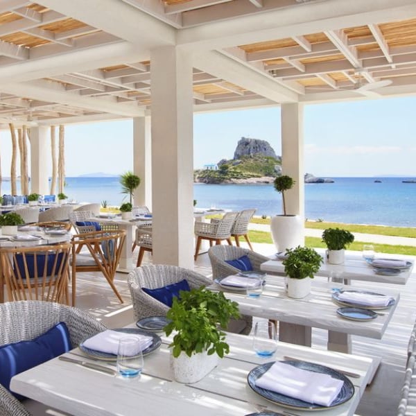 Outdoor dining area at Ikos Aria with tables, greenery, and a view of the coast and a rocky islet with a white chapel.