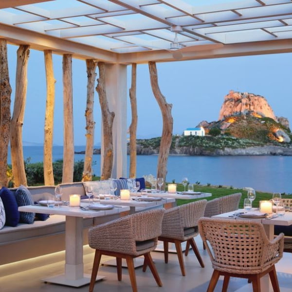Outdoor dining area at Ikos Aria with candle-lit tables and wicker chairs, overlooking a scenic island with a church on a rocky hill.