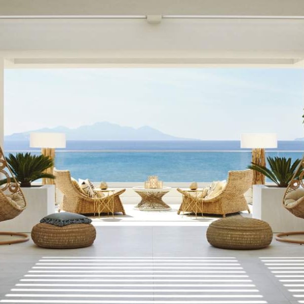 Veranda at Ikos Aria with hanging chairs, wicker armchairs, and a coffee table overlooking the sea and distant mountains.