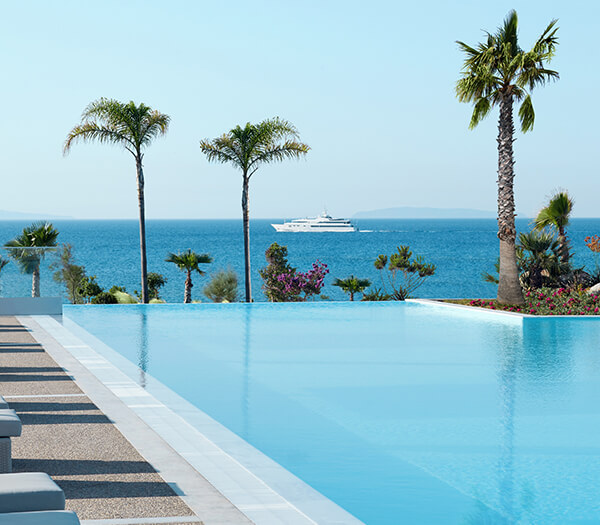 Infinity pool at Ikos Aria with palm trees and a yacht in the Aegean Sea background.