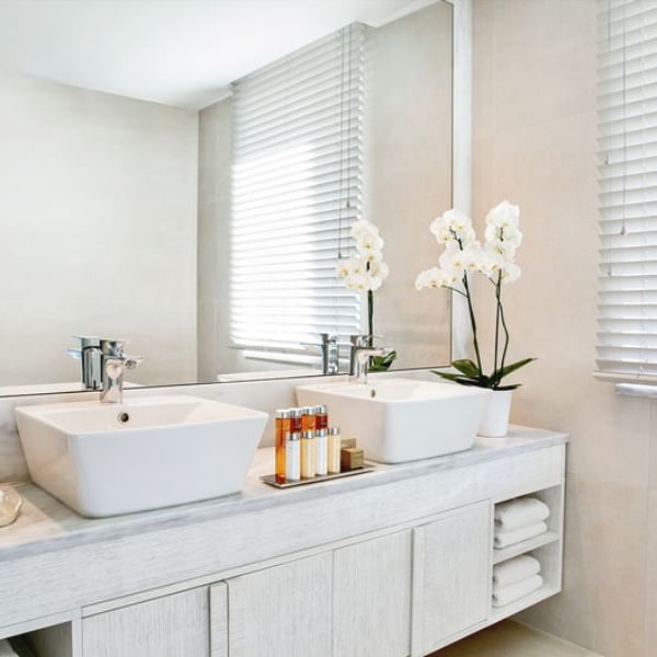 Luxurious bathroom at Ikos Aria with dual sinks, white orchids, toiletries, and a large window with blinds.
