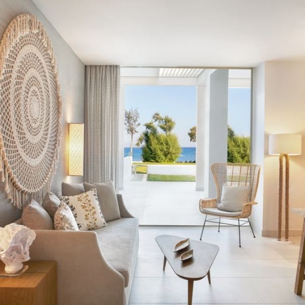 Ikos Aria living room with a sofa, decorative cushions, woven wall hanging, wooden table and chair, and large windows with sea views.