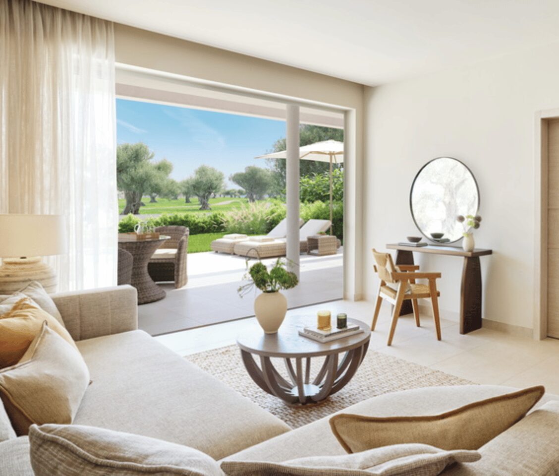 Living room of Ikos Odisia Deluxe Two Bedroom Bungalow Suite with garden view, featuring a comfortable sofa, coffee table, and access to a private garden with outdoor seating.