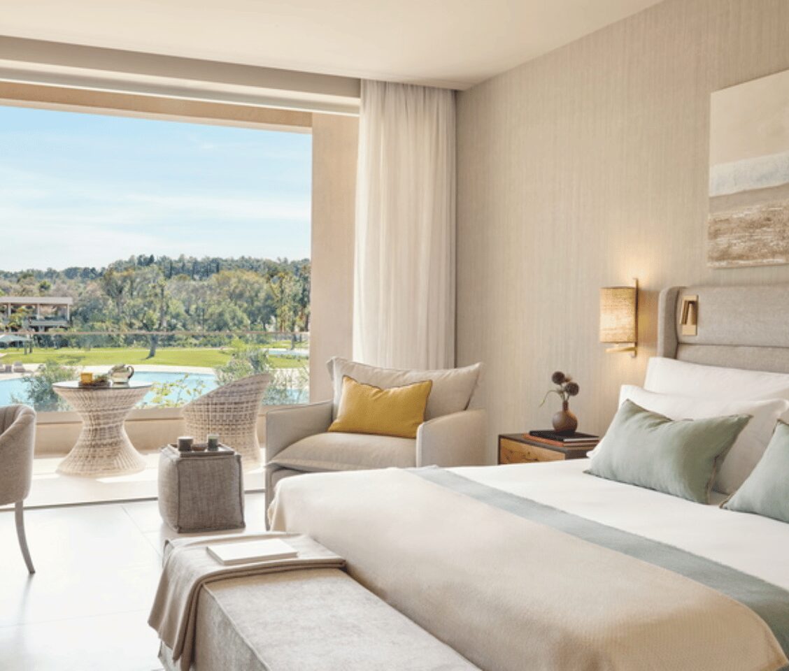 Family room at Ikos Odisia with beige decor, large bed, seating area, and a view of the pool and greenery through a window.