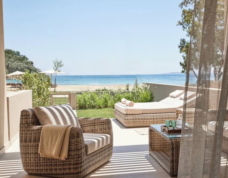 View from Ikos Odisia balcony showing wicker furniture, a lush garden, and a scenic beach with a blue sea backdrop.