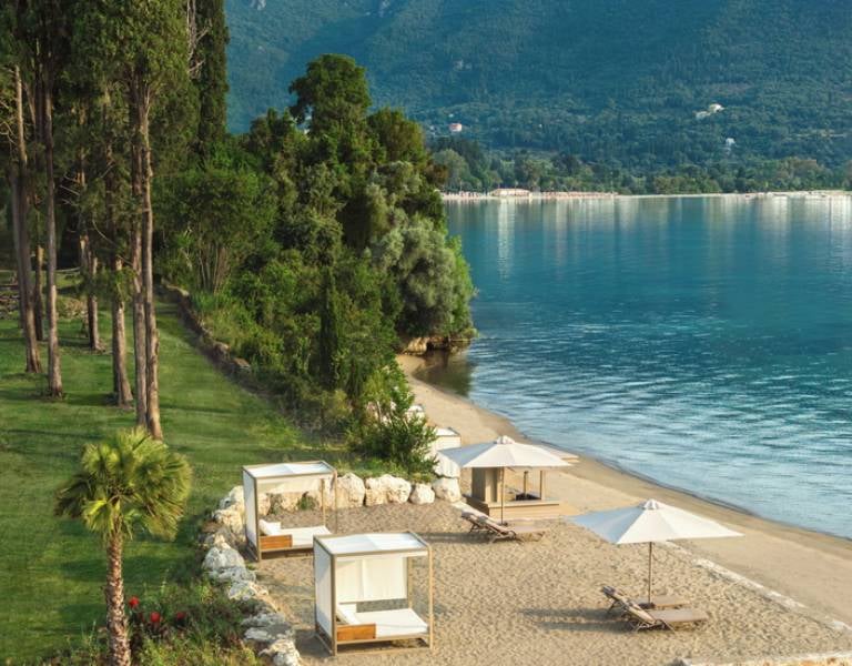 Ikos Odisia Beach with sun loungers and umbrellas, lush greenery, and mountains in the background.