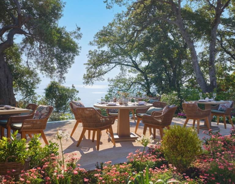 Outdoor dining area at Ikos Odisia Gallery with wicker chairs, wooden tables, surrounded by greenery and a sea view.
