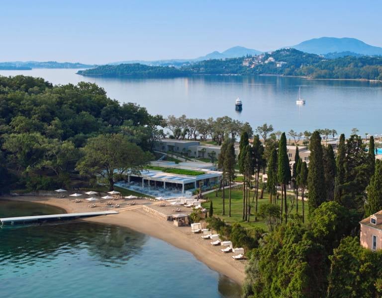 Aerial view of Ikos Odisia resort with sandy beach, sun loungers, green gardens, and a beachfront pavilion nestled in a coastal bay.