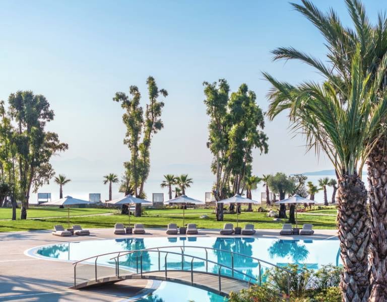 Pool area at Ikos Odisia resort with palm trees, sun loungers, and a view of the beach and sea.