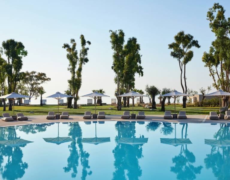 Outdoor pool at Ikos Odisia with surrounding trees, white parasols, and the sea in the background.