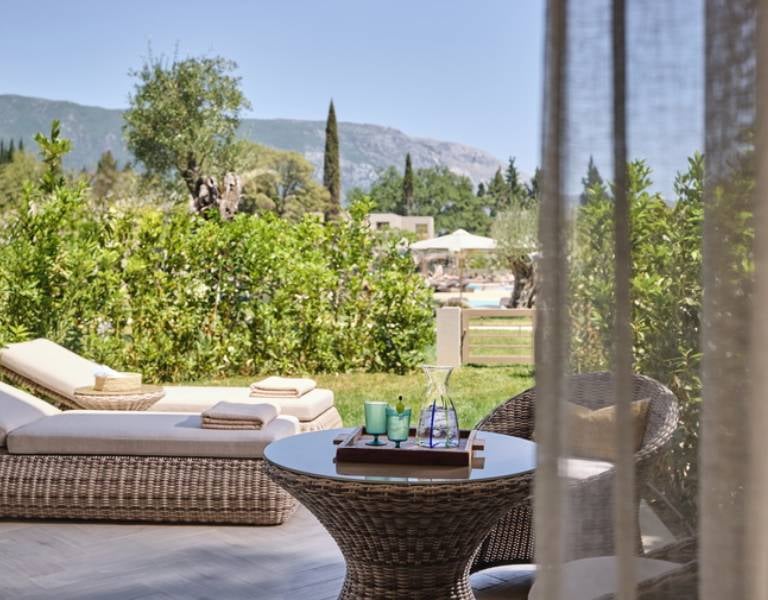 Private garden at Ikos Odisia with wicker lounge chair and table, surrounded by greenery and mountain views in the background.