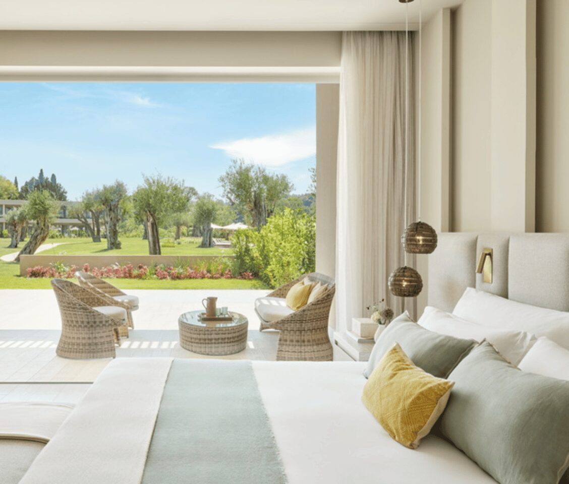 Interior of an Ikos Odisia one-bedroom suite with a private garden view, showcasing a spacious bed, soft pillows, and outdoor seating overlooking a lush landscape.