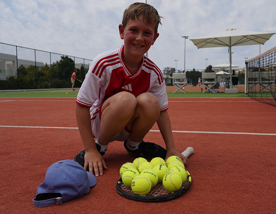 Young boy kneeling on a tennis court with a tennis racket and yellow balls, wearing a red and white jersey, smiling at the camera.